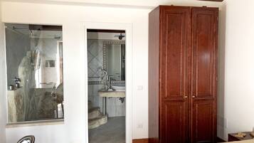 A bedroom with a bed, a wooden wardrobe, and a bathroom with a shower, sink, and mirror.
