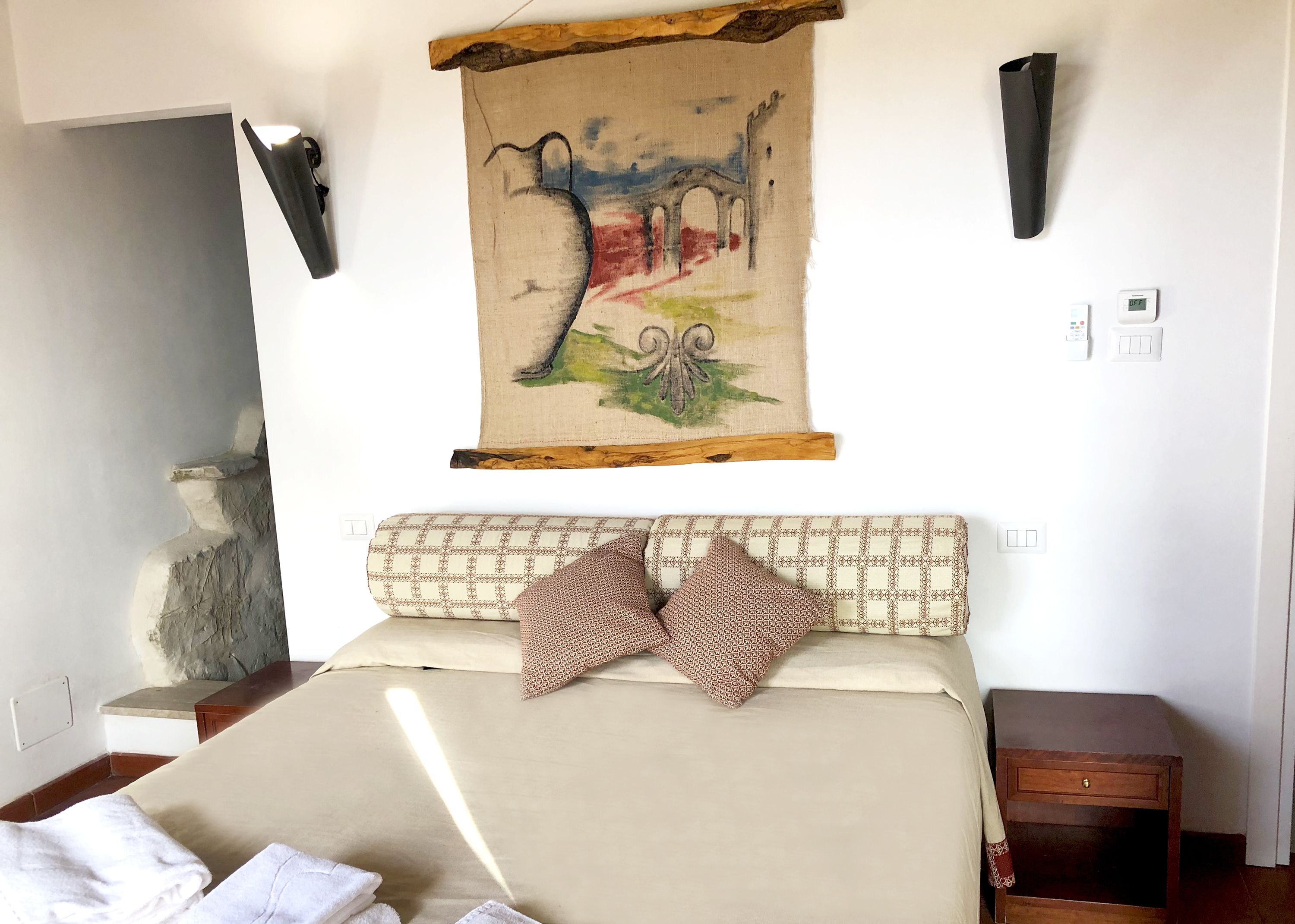 A neatly made bed with a patterned headboard, a wall-mounted tapestry, and a small wooden nightstand.