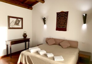 A neatly made bed with a beige bedspread and pillows, a wooden nightstand, a small table with a lamp, and wall-mounted lights. - Resort Fonte Vulci (Canino)