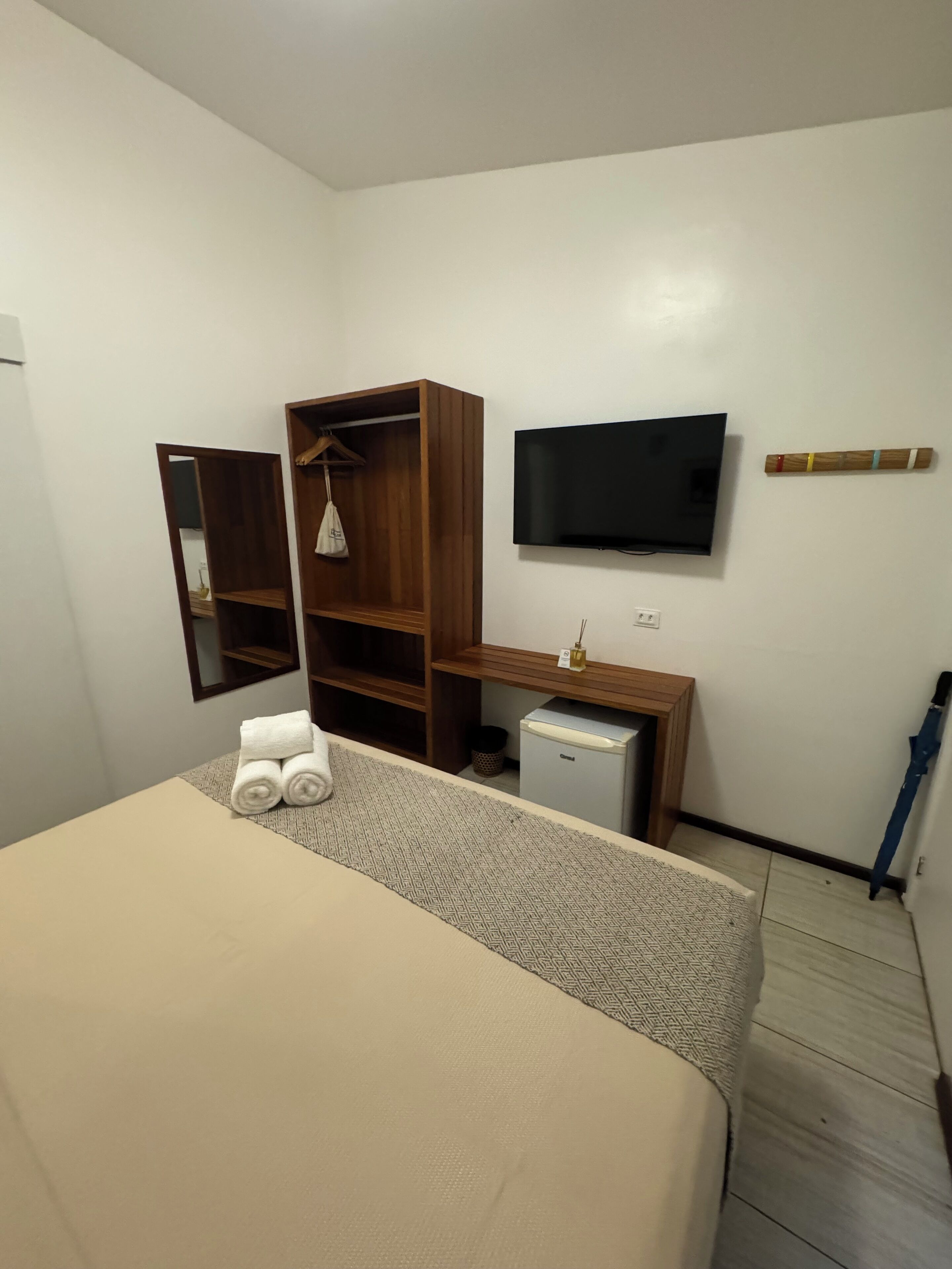 A hotel room with a bed, a wooden wardrobe, a small fridge, a television, and a wall-mounted towel rack.