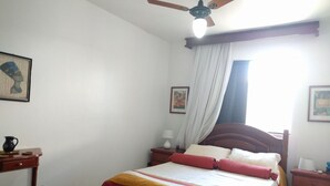 A bedroom with a bed, a nightstand, a bedside table, a ceiling fan, and a window with curtains. - Habitat Villa Florida Praia Ingleses (Florianópolis)
