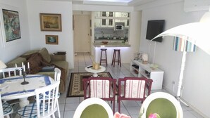 A living room with a dining area, a kitchen in the background, and a TV mounted on the wall. - Habitat Villa Florida Praia Ingleses (Florianópolis)