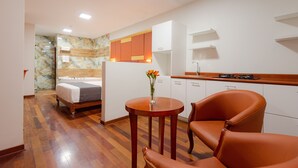 Ein kompaktes Hotelzimmer mit Bett, einem kleinen runden Tisch mit Vase, einem braunen Ledersessel, einer Küchenzeile und einem Badezimmer mit Marmorwand.