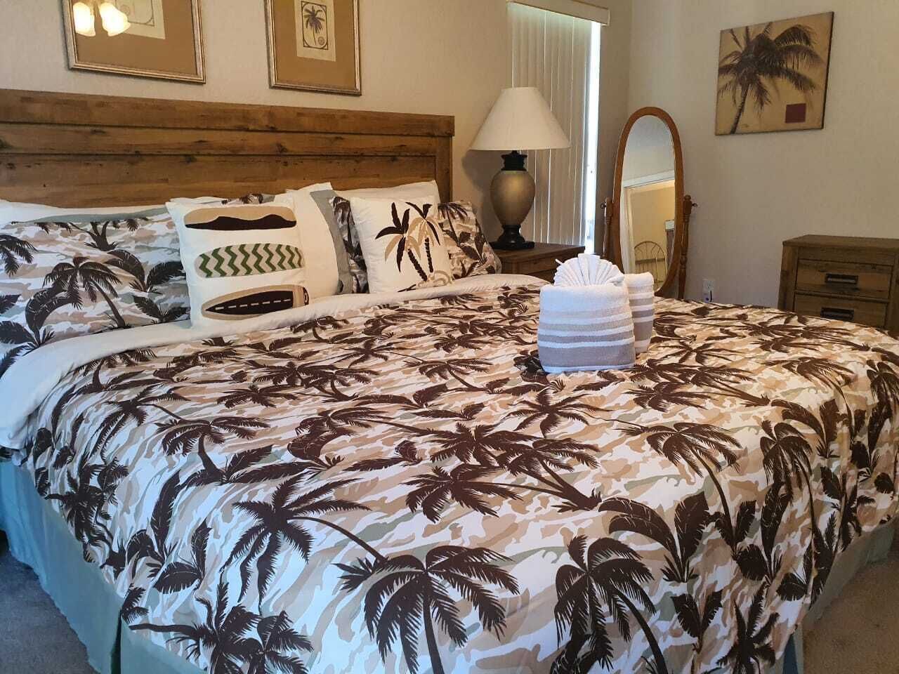 Une chambre à coucher avec un lit orné d’une couvre-lit à motifs de feuilles de palmier tropicales, une tête de lit en bois, une lampe de chevet, un miroir et une table de chevet en bois.