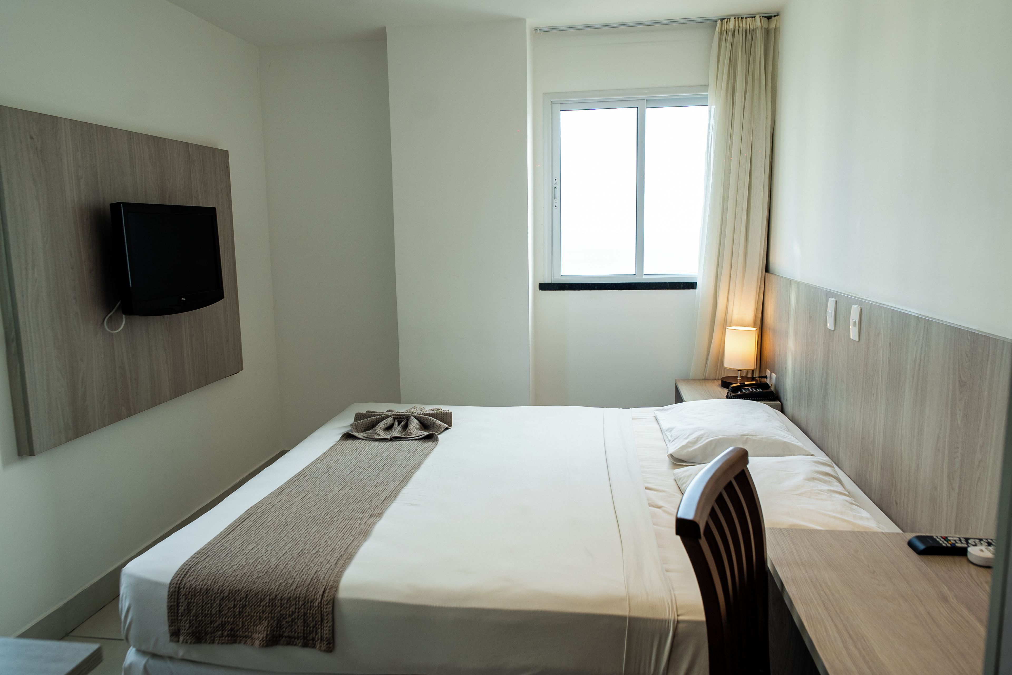 A hotel room with a bed, a bedside table with a lamp, a TV mounted on the wall, and a window with curtains.