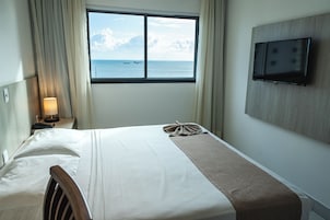 Ein Hotelzimmer mit einem Bett, einem an der Wand montierten Fernseher, einem Fenster mit Meerblick und einer Lampe auf dem Nachttisch.
