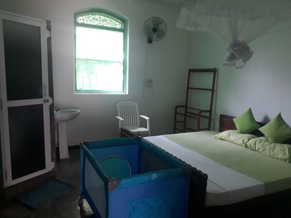 A bedroom with a bed, two chairs, a window, a fan, and a small bathroom area. - Villa Lagoonedge (Dodanduwa)