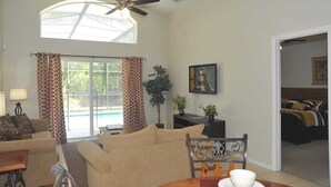 A living room with a ceiling fan, a sofa, a dining table, and a view of a pool through large windows.