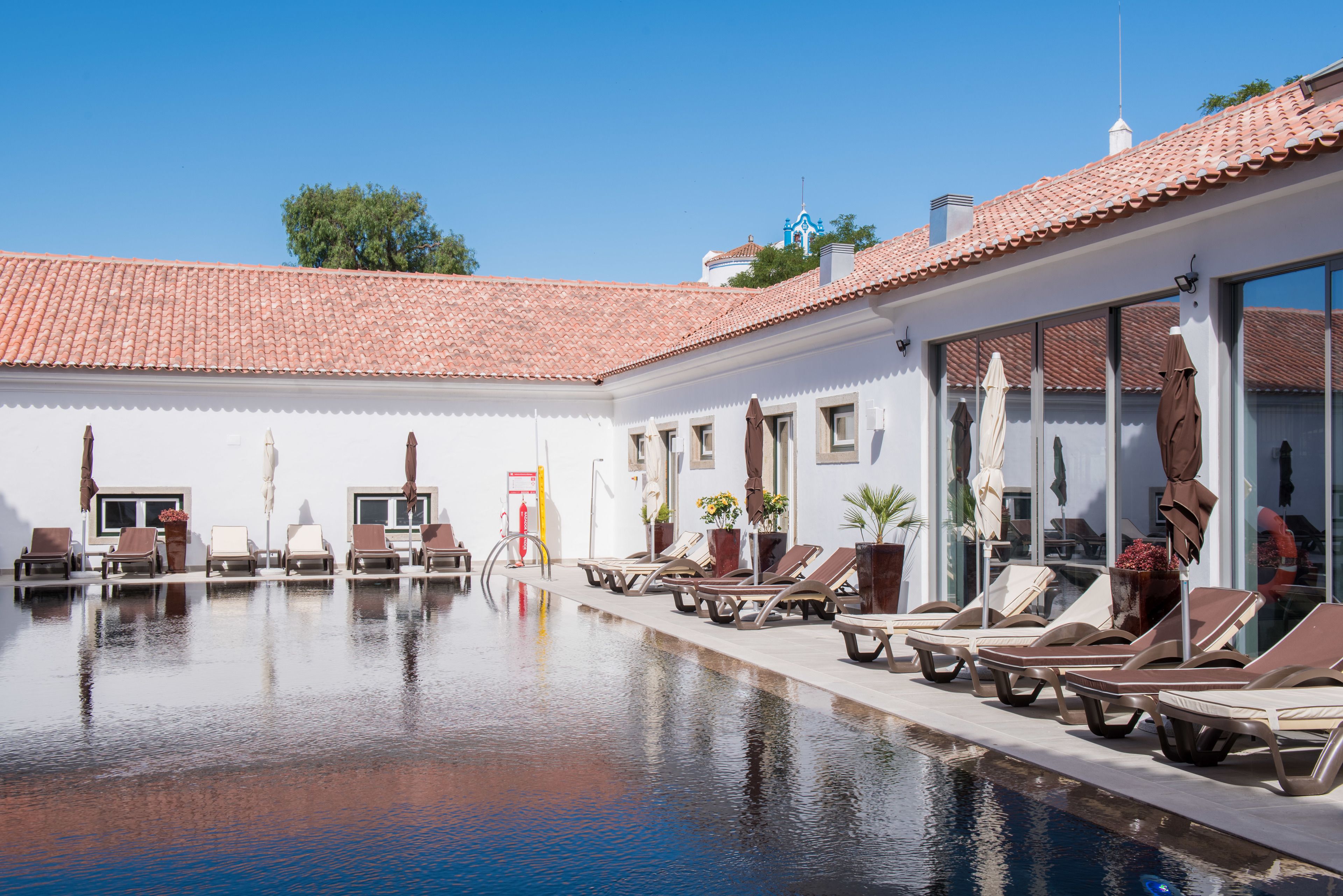 Zona de piscina con sillones de descanso y sombrillas, techo de baldosas y cielo despejado.