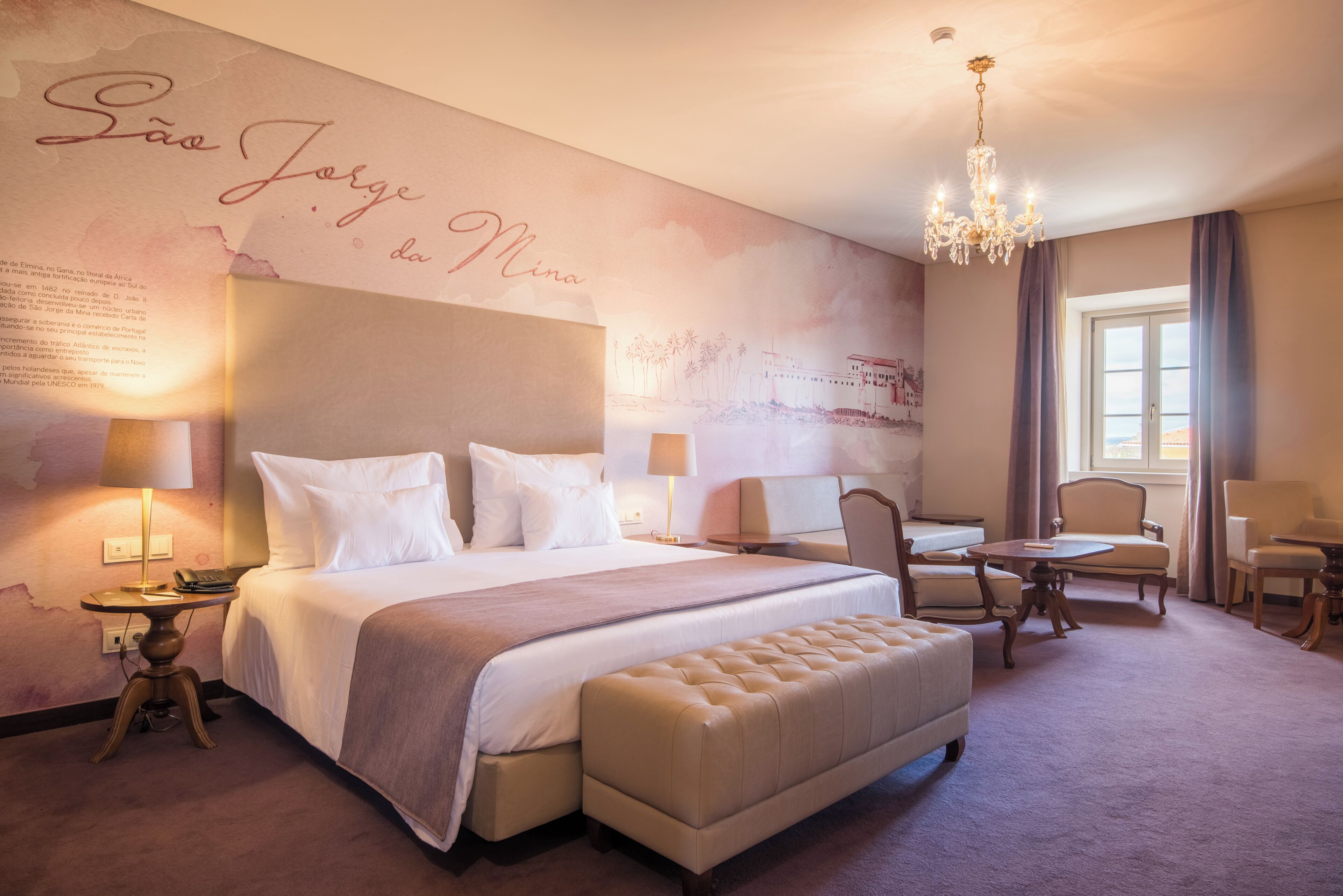 A hotel room with a large bed, a sitting area with a table and chairs, a chandelier, and a wall with text and a map.