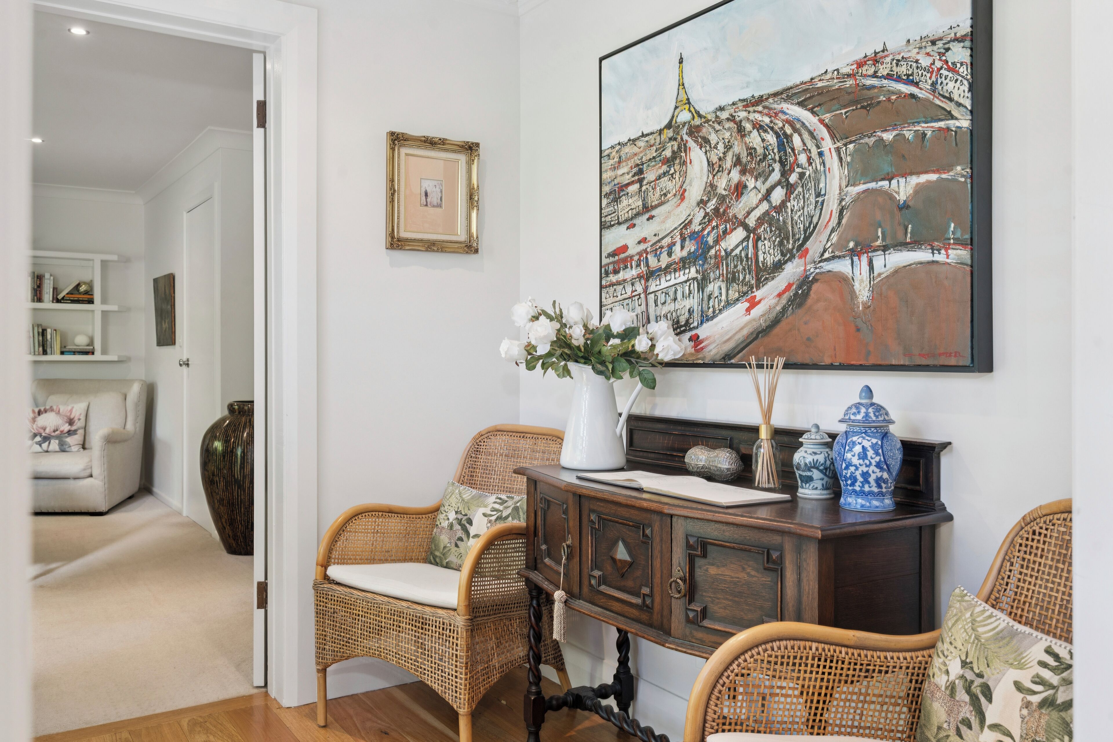 Una habitación con sillas de mimbre, un armario de madera y un cuadro grande con una vista urbana.