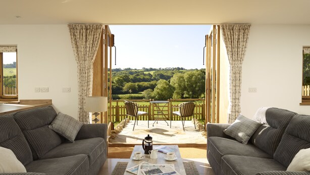 Un salon avec un canapé et des fauteuils, une table basse avec des journaux, et une vue sur un paysage verdoyant par une porte ouverte.