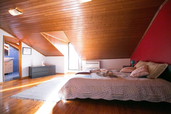 Quarto com teto inclinado, piso de madeira, uma cama com colcha branca, mesa de cabeceira e cômoda.