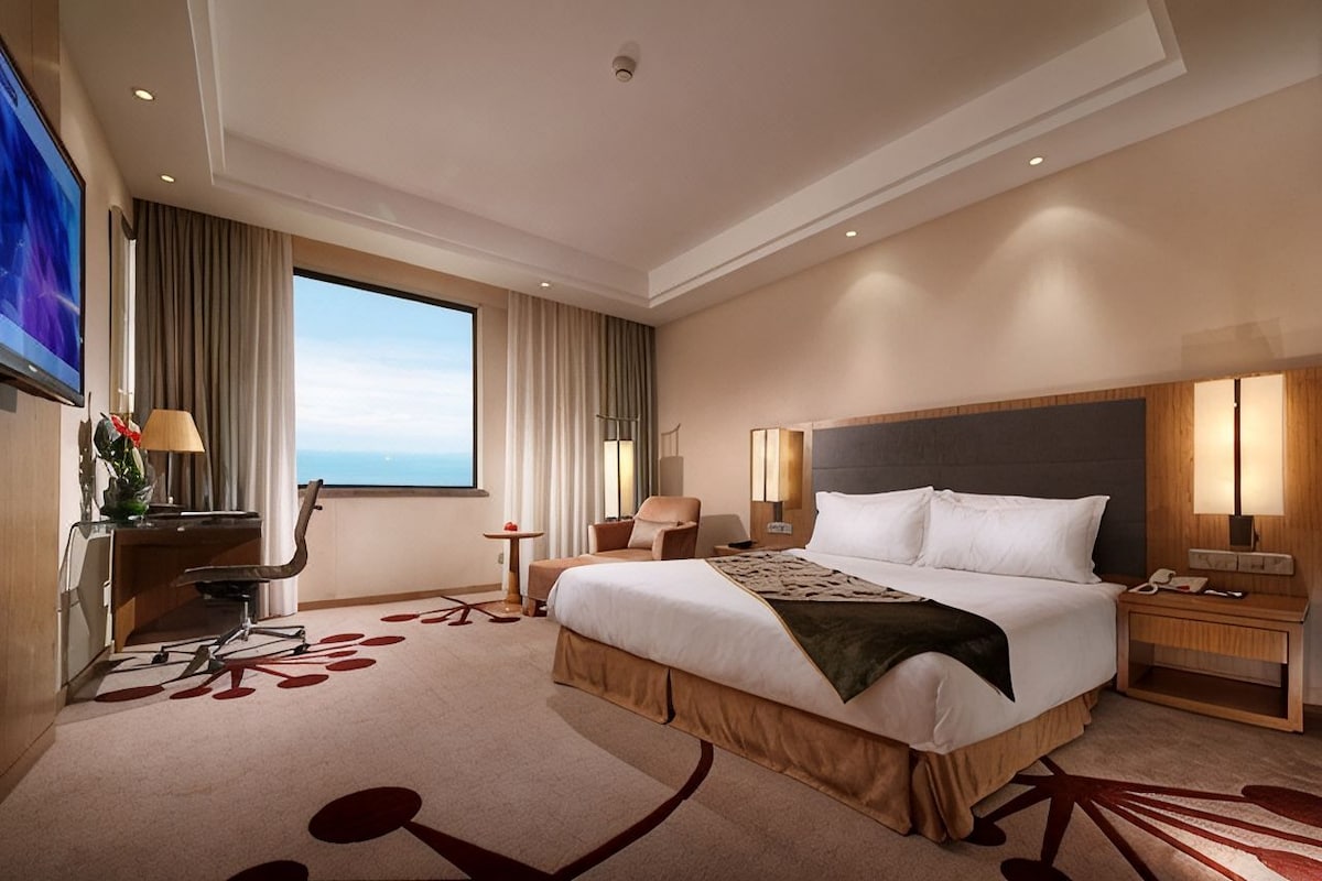 Una camera d'albergo con un letto grande, una scrivania con una sedia, una TV e vista sul cielo.