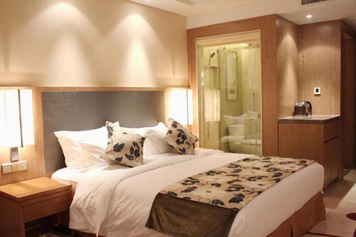 Una camera d'albergo con un letto, lampade sui comodini, una testiera in legno, un armadio e un bagno con porta in vetro.