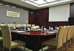 Un salón de conferencias con una mesa larga, sillas, micrófonos, botellas de agua y una pantalla de proyector.