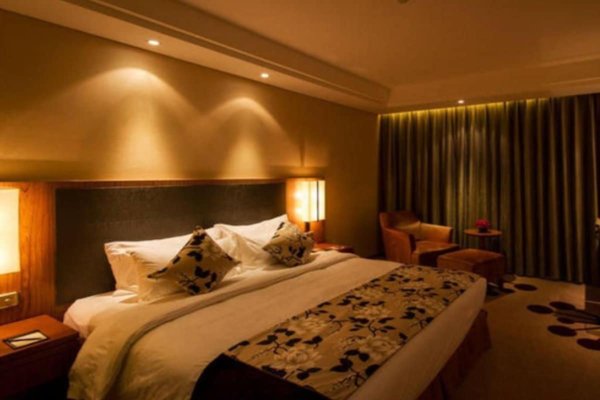 Camera d'albergo con un letto grande, due lampade ai comodini, una sedia, un tavolino e vista all'esterno tramite tende.