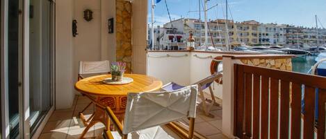 Un balcón con mesa y sillas de madera, una planta en maceta y vistas a un puerto deportivo con barcos y edificios al fondo.