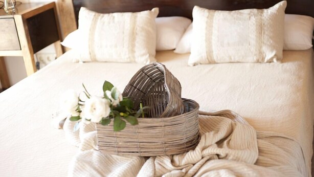 Un lit bien fait, avec du linge de lit et des oreillers blancs, un panier en osier rempli de fleurs et une table de chevet en bois sur laquelle est posé un vase de fleurs.