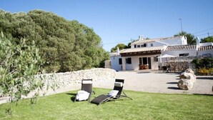 Une villa avec un mur en pierre, un patio doté de deux chaises longues et, au fond, un bâtiment blanc avec un toit en tuiles.