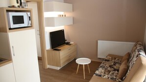 Un espacio habitable pequeño con kitchenette, televisor, un sofá blanco y un taburete blanco.