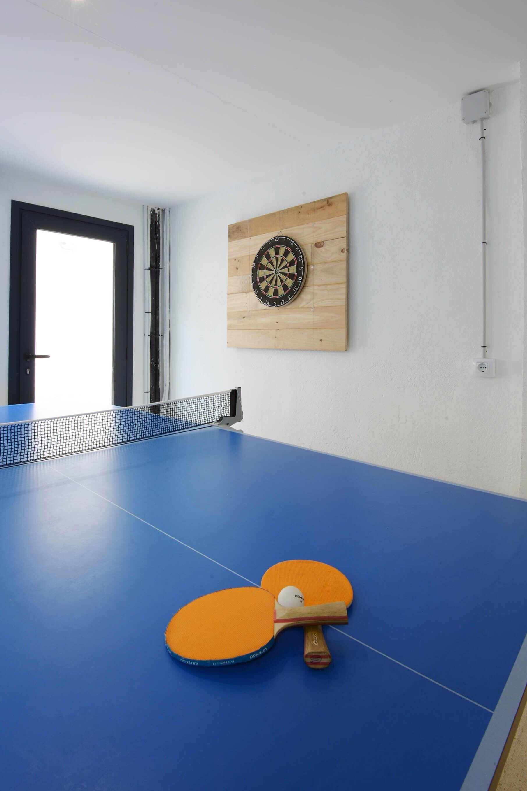 Een tafeltennistafel met net, een dartbord en twee tafeltennisrackets op een blauwe vloer.