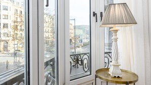 Ett rum med utsikt över en stadsgata genom ett stort fönster, ett litet runt bord med en vit lampa och ett balkongräcke.