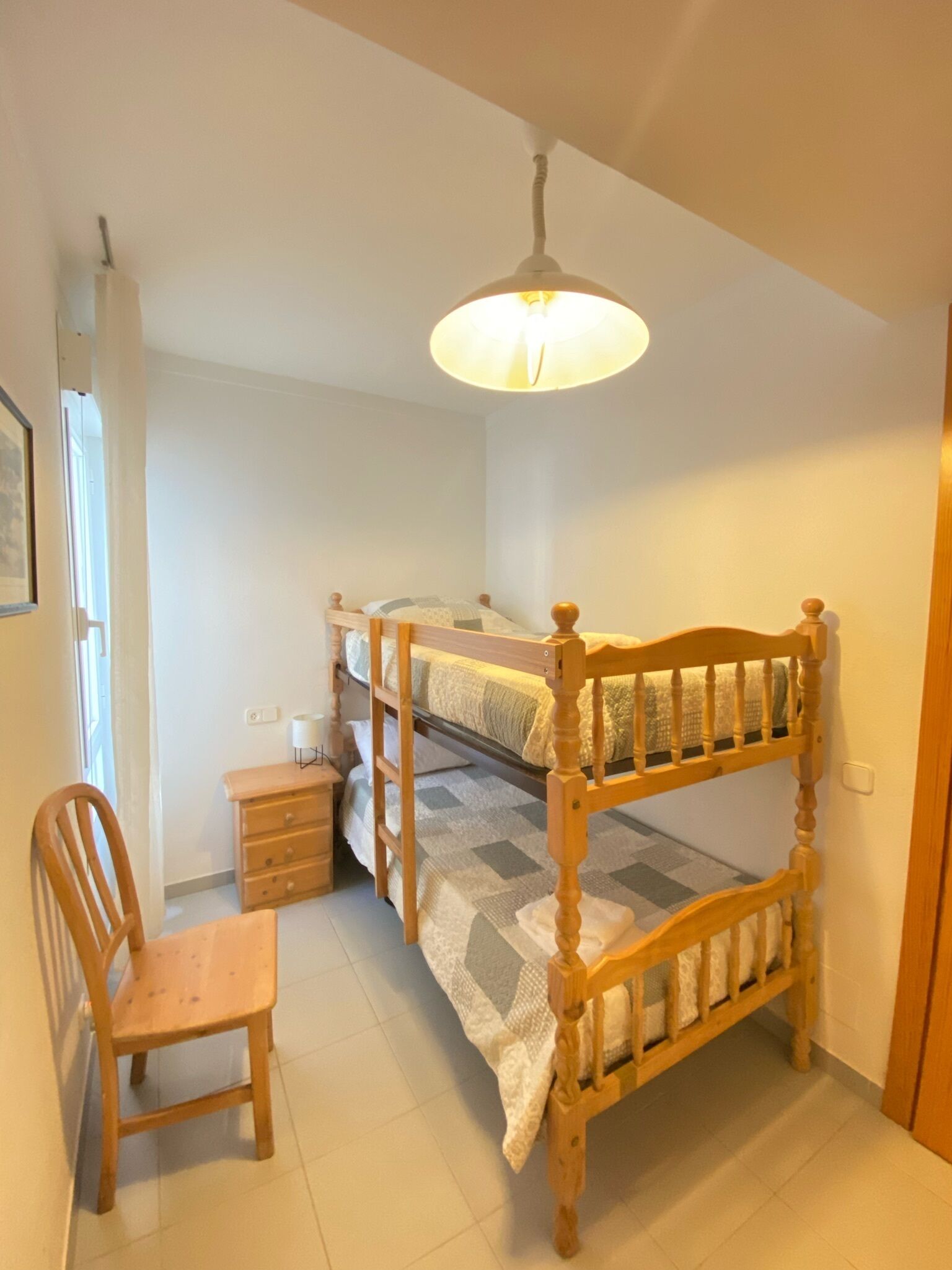 Une petite pièce chaleureuse, avec un lit superposé, une chaise en bois, une table de chevet et un luminaire suspendu.