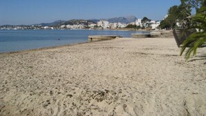 Une plage de sable fin, un ciel bleu azur, une mer calme et, au loin, une vue sur la ville avec ses bâtiments et ses montagnes.