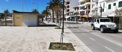 Un sendero pavimentado con un pequeño quiosco, un edificio blanco con balcones y una camioneta SUV blanca estacionada en la calle.