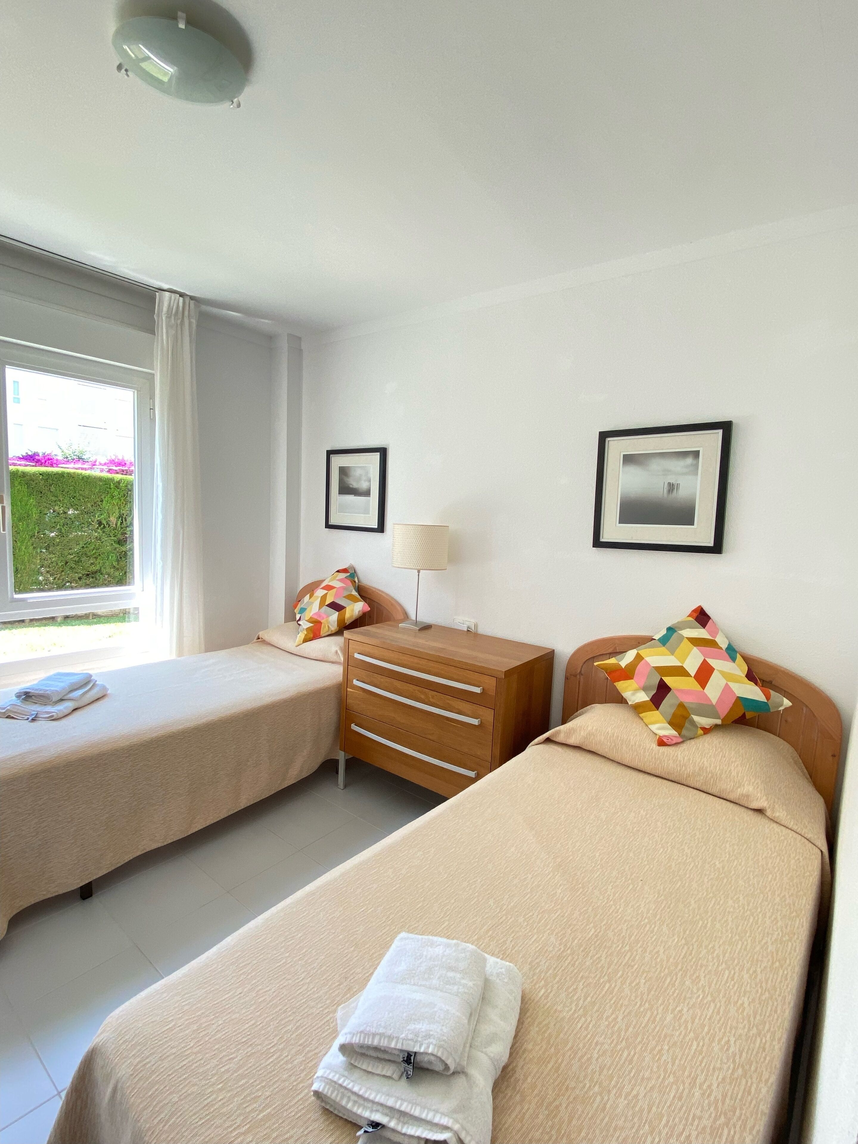 A hotel room with two beds, a wooden dresser, a lamp, and framed pictures on the wall.