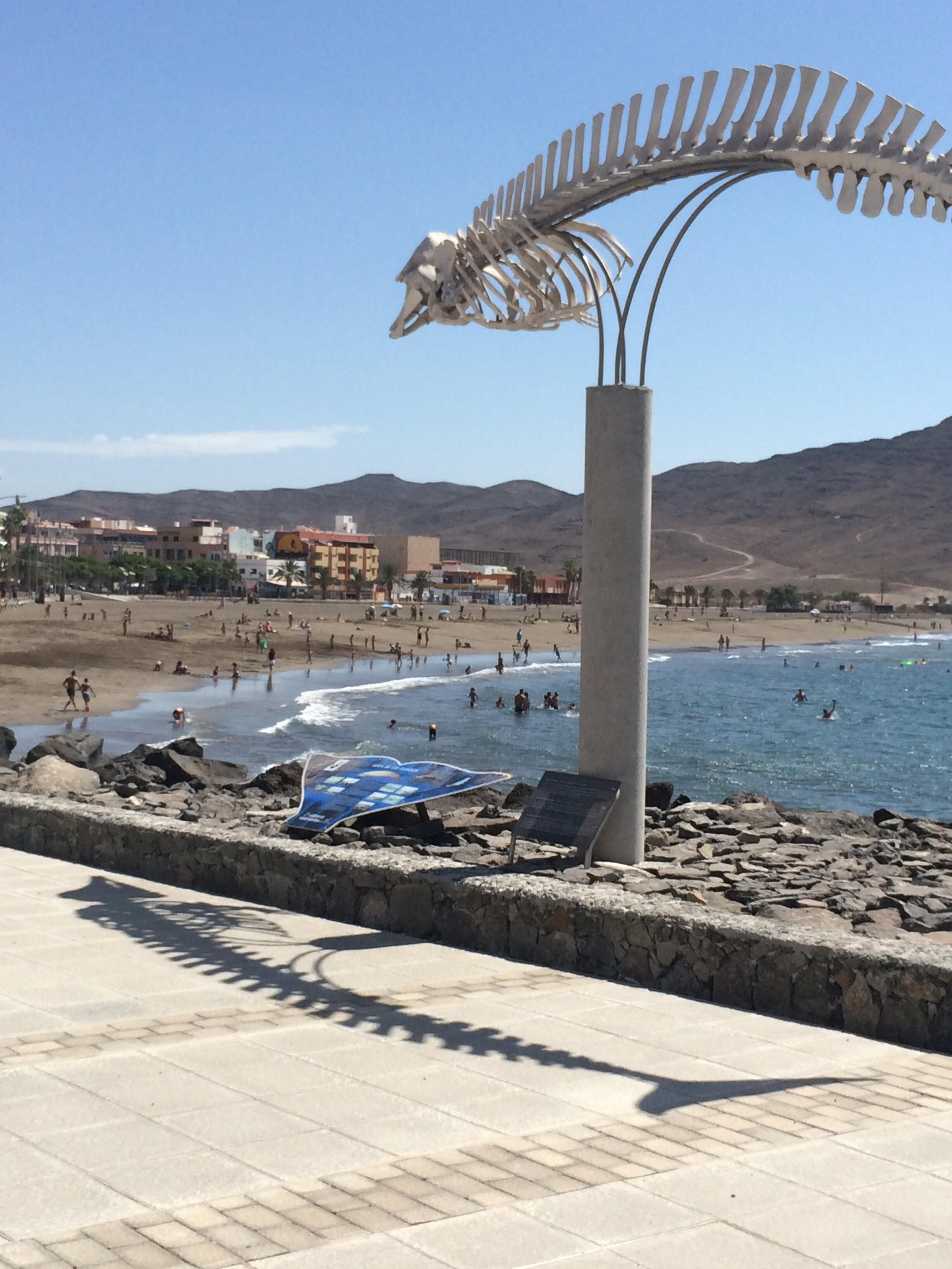 A beach with a large metal sculpture, rocky shoreline, and buildings in the background.