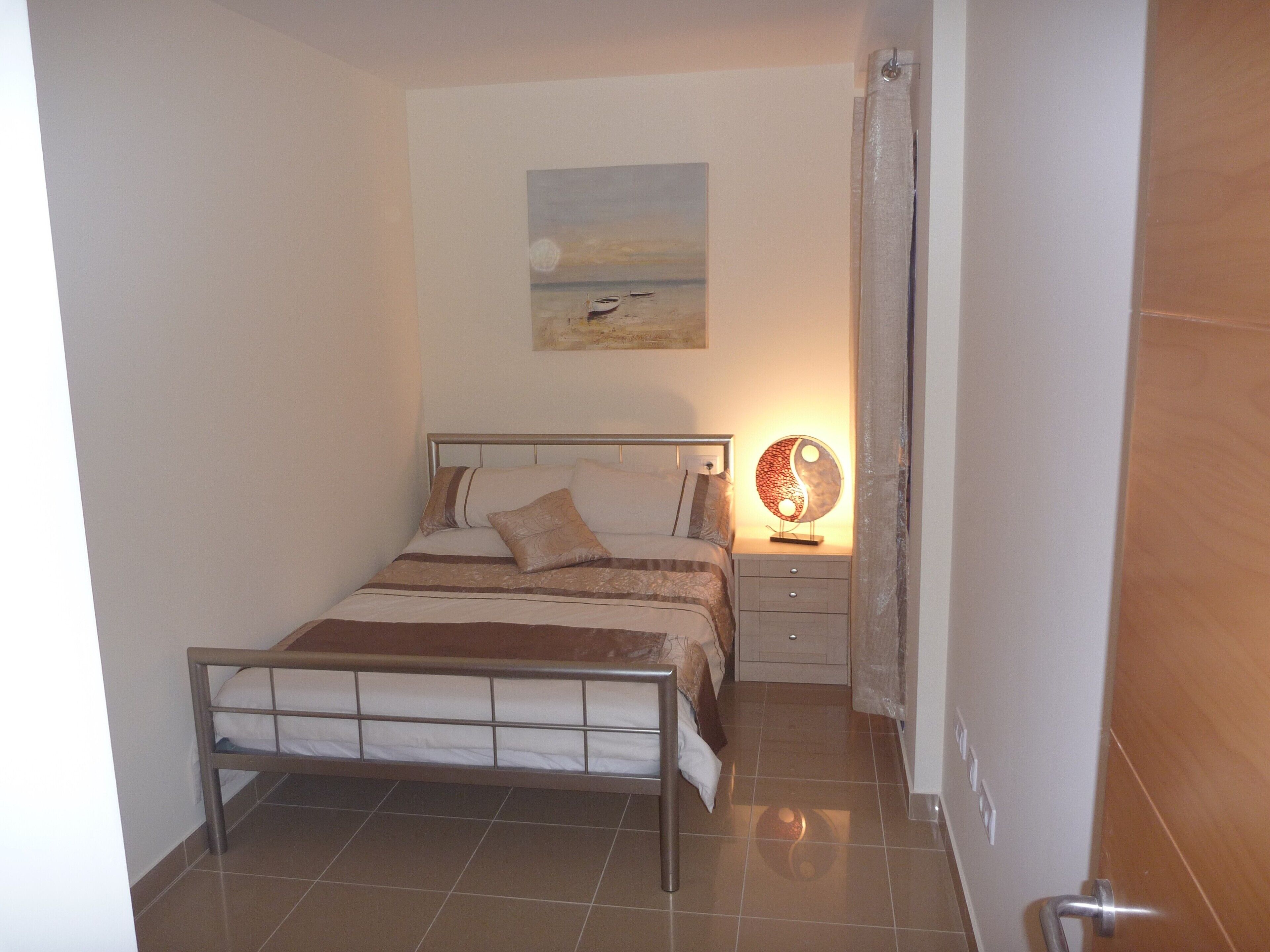 Un dormitori amb llit, taula de nit, làmpada i un quadre a la paret.