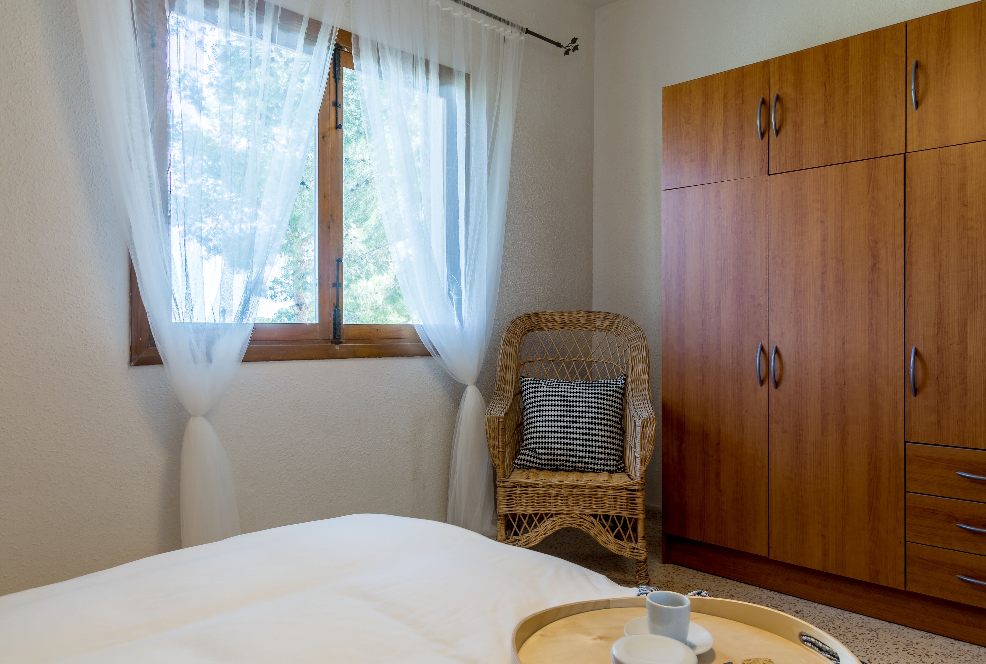Une chambre à coucher avec un lit, une chaise en osier, une armoire en bois et une fenêtre ornée de rideaux légers.