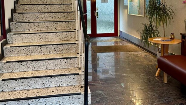 A staircase with a glass railing leading to a high ceiling, a red door at the bottom, and a hallway with a polished floor and a round table with a vase.