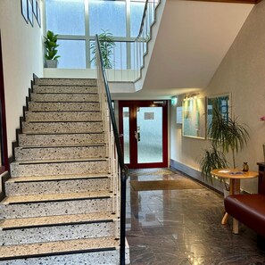 Escadaria com guarda-corrimão de vidro que leva a um teto alto, porta vermelha na base e corredor com piso polido e mesa redonda com um vaso.