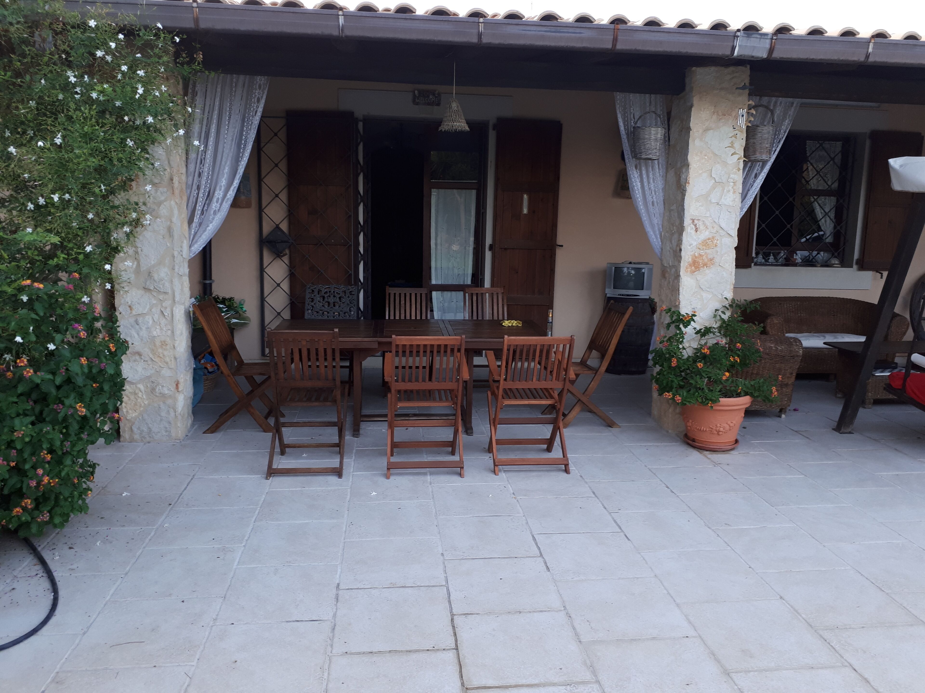 Eine Terrasse im Freien mit Holztisch und -stühlen, einem Fernseher und Topfpflanzen.