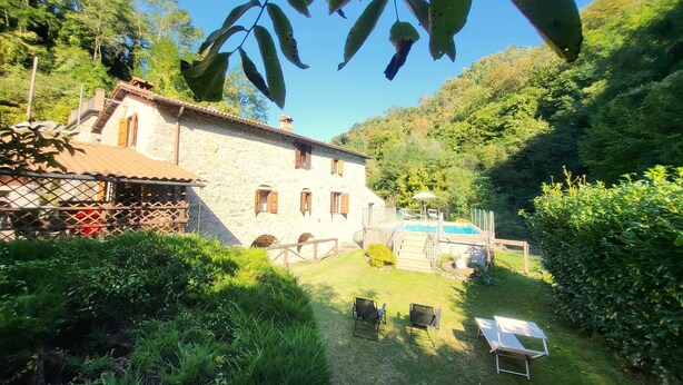 A stone house with a swimming pool, surrounded by lush greenery and a clear blue sky.