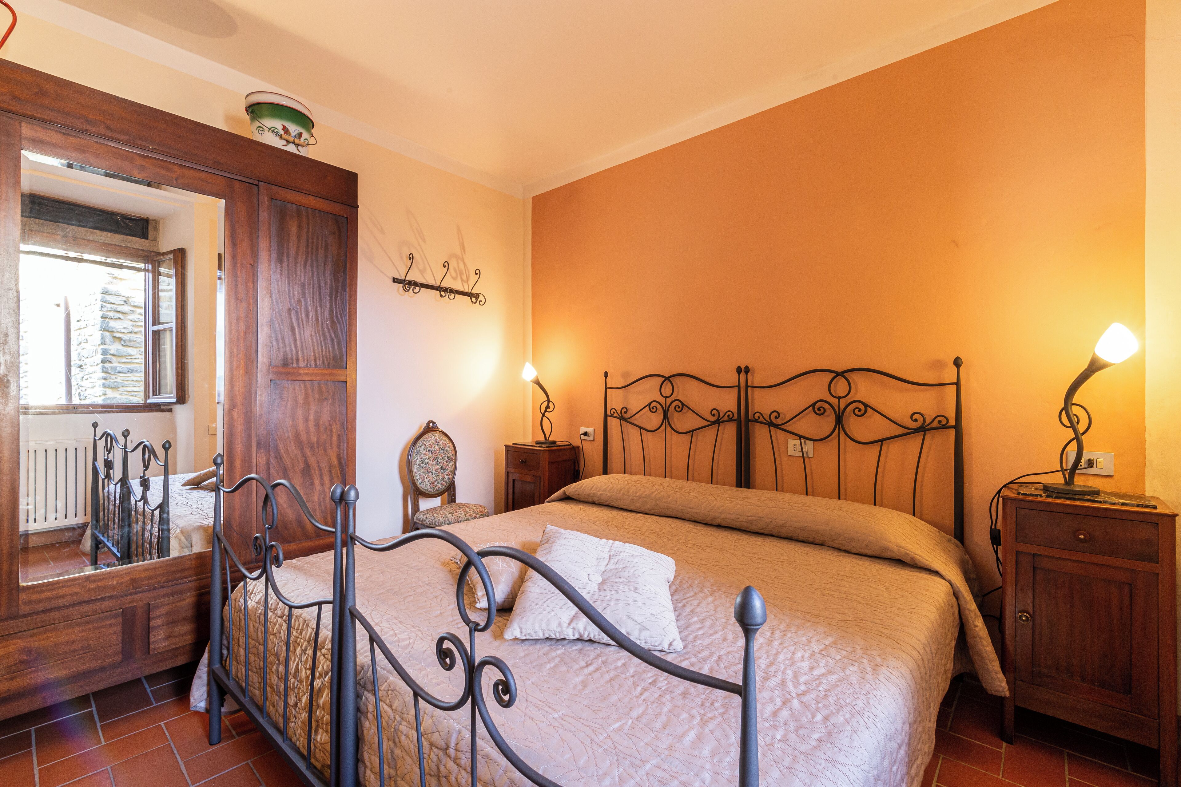 A bedroom with a metal bed frame, a wooden wardrobe, a nightstand with a lamp, and a mirror.