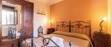 A bedroom with a metal bed frame, a wooden wardrobe, a nightstand with a lamp, and a mirror.