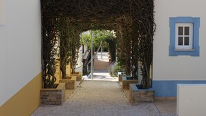 A pathway leading through a covered archway with plants on either side, flanked by low walls and a small window.