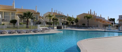 A swimming pool with lounge chairs and umbrellas, surrounded by buildings with tiled roofs and chimneys.