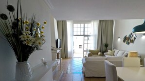 A modern living room with a sofa, a dining table, and a TV. There is a vase with flowers on the wall and a small stool.