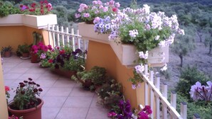 Un balcone con piante e fiori in vaso, ringhiera bianca e vista su un paesaggio montuoso.