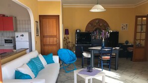 Un soggiorno con un divano bianco, una poltrona blu, un tavolo da pranzo con sedie, un angolo cottura con frigorifero e una porta in legno.