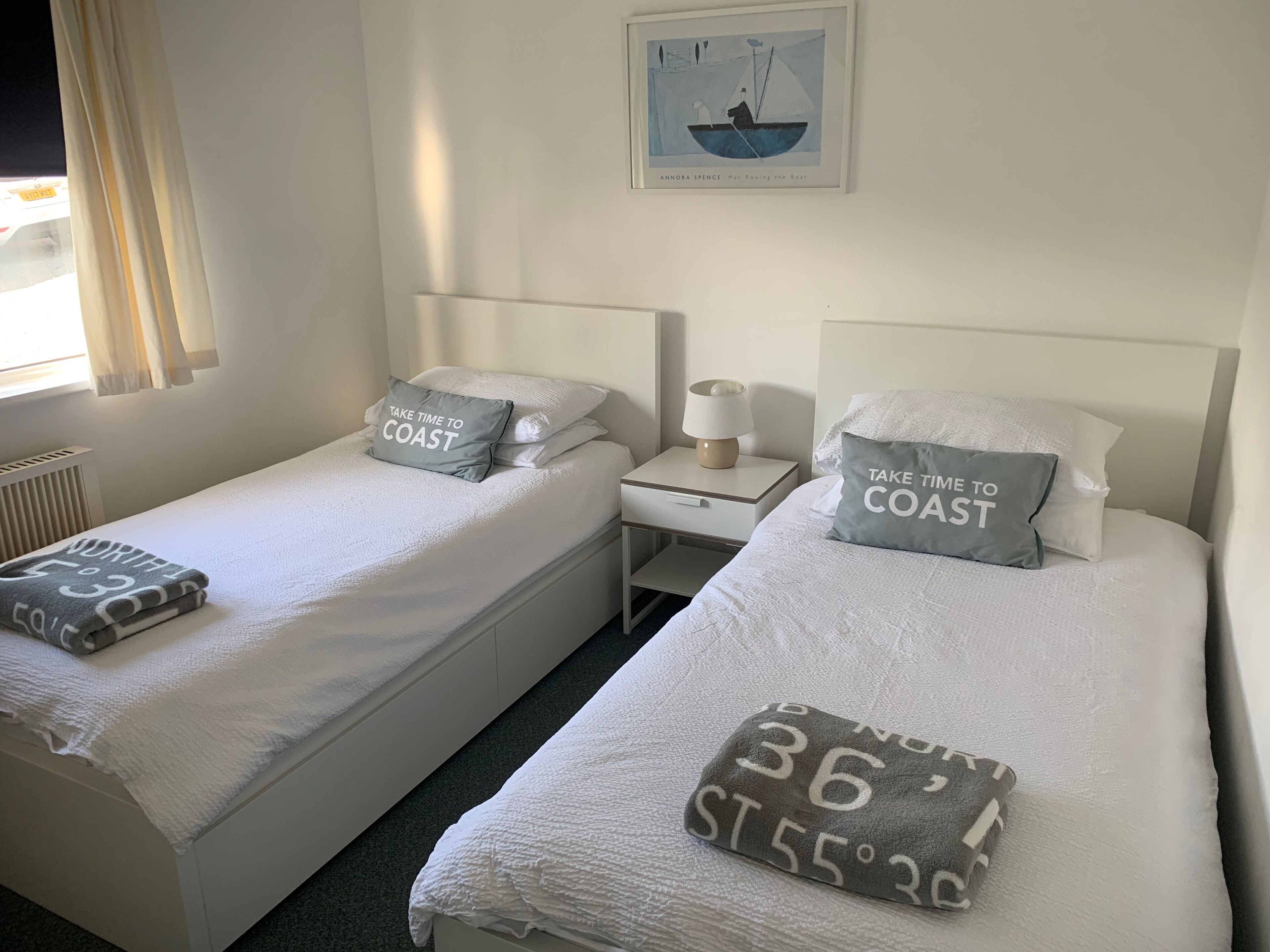 A hotel room with two single beds, each with a pillow that says "TAKE TIME TO COAST." There is a framed picture of a sailboat on the wall, a bedside table with a lamp, and a window with curtains.