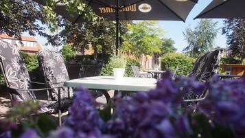 An outdoor seating area with tables and chairs, surrounded by greenery and a clear sky.