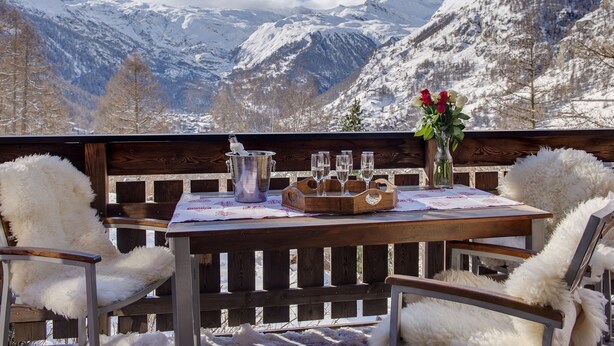 A wooden table with a metal bucket, glasses, and a vase with flowers, set against a snowy mountain backdrop.