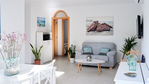 Una sala de estar moderna con un sofá gris, sillas blancas, una mesita redonda y una cocina con nevera y armarios.