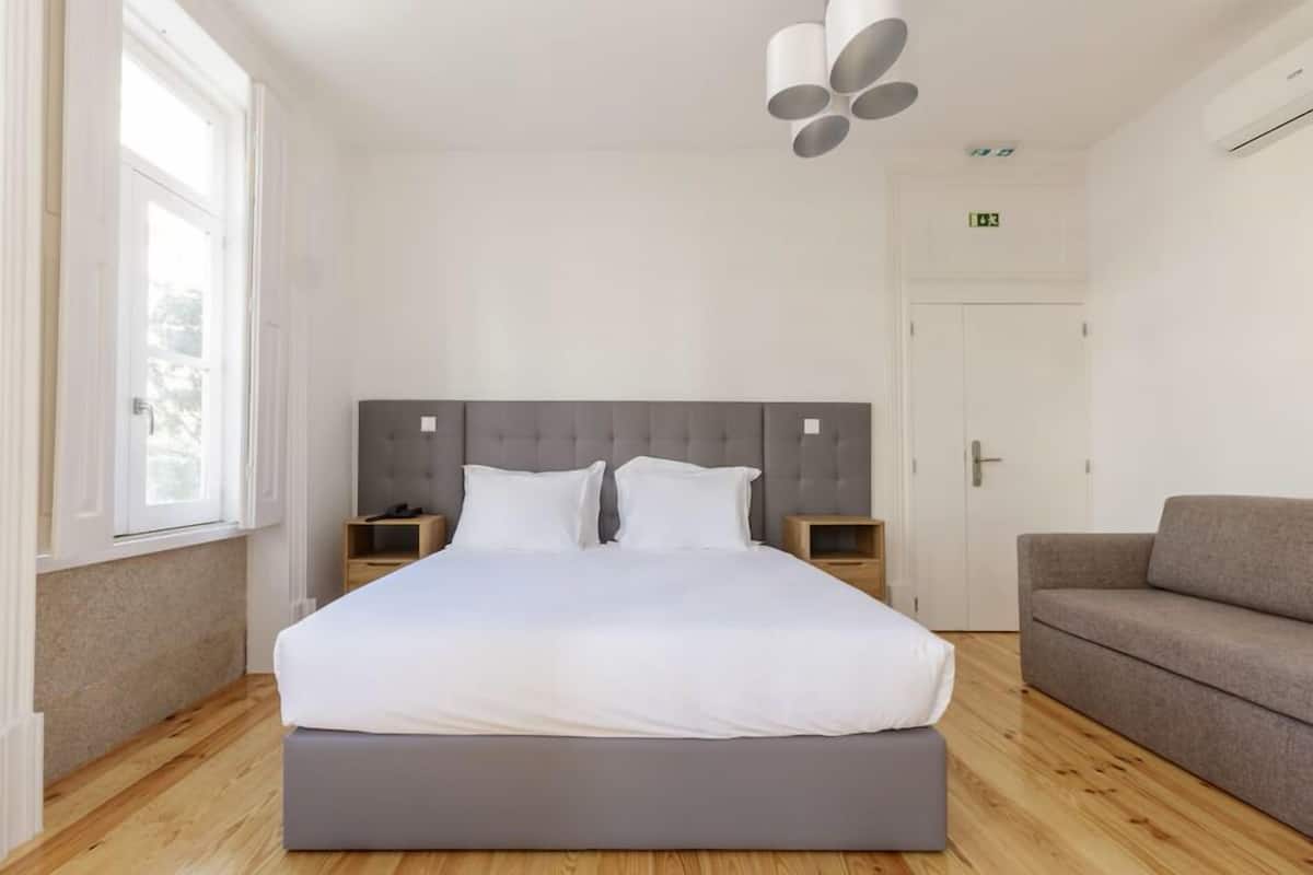 A modern bedroom with a large bed, a grey upholstered headboard, bedside tables, a grey sofa, and a window providing natural light.